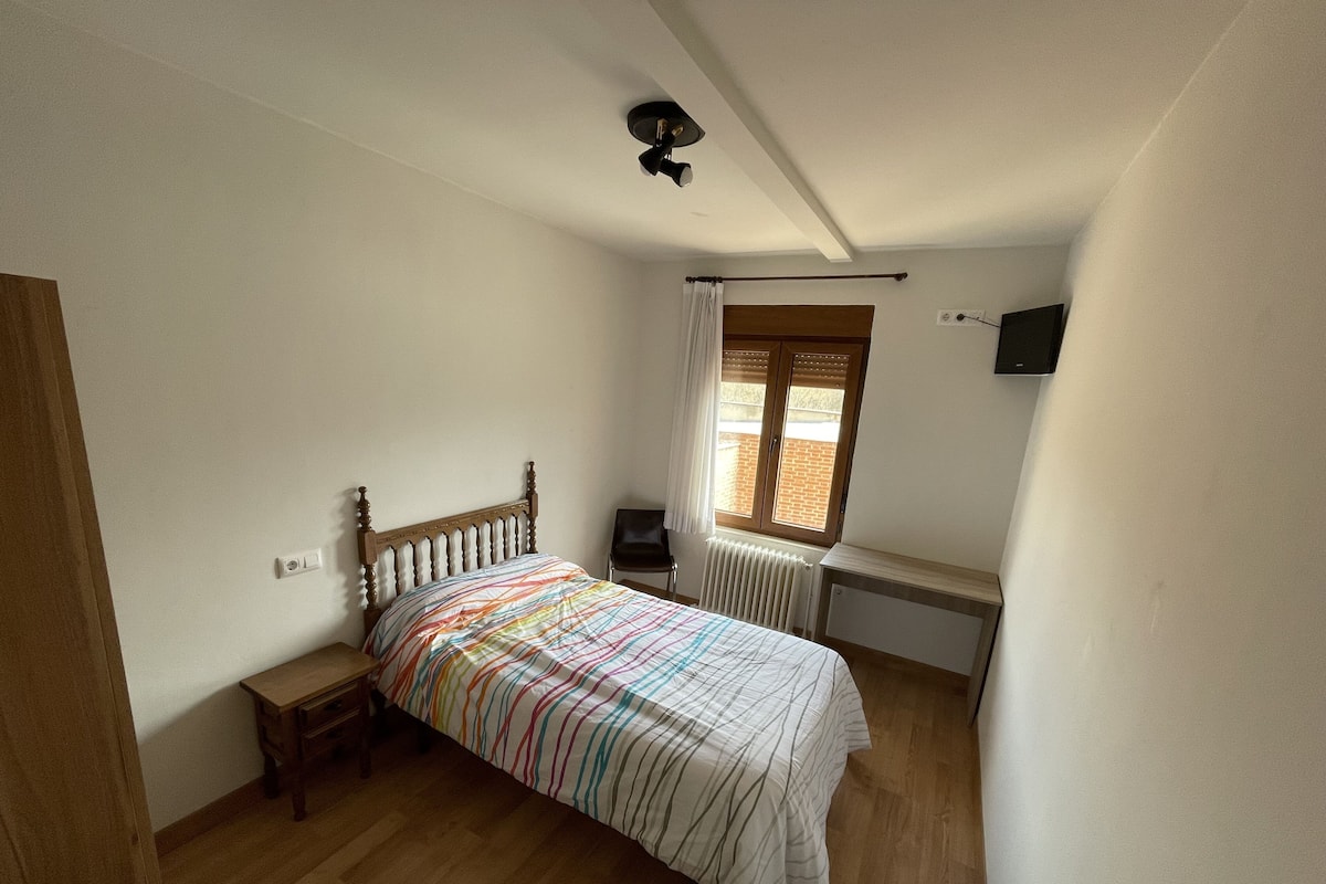 A bedroom with a wooden bed, a nightstand, a desk, a radiator, and a window with curtains.