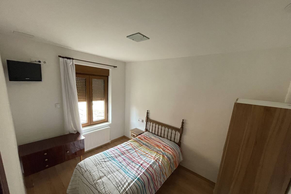 A bedroom with a bed, a window with curtains, a bedside table, and a TV mounted on the wall.