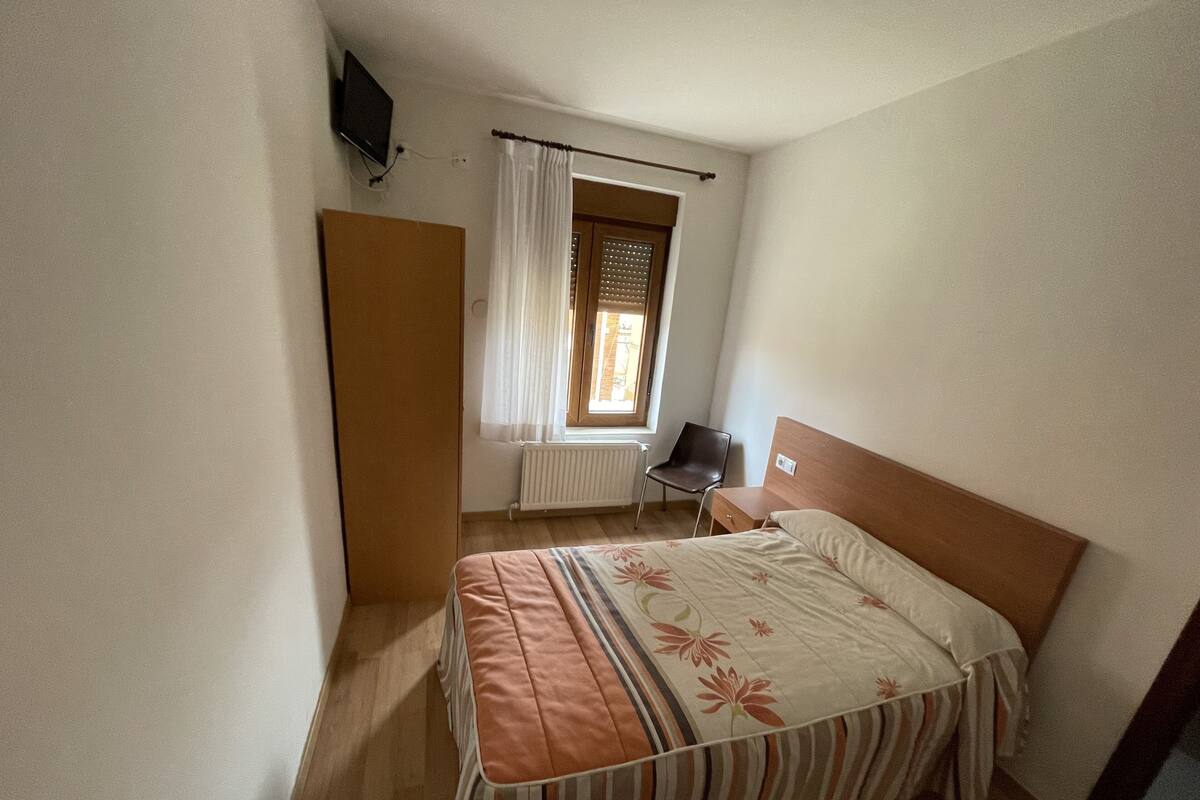 A bedroom with a bed, a chair, a window with curtains, and a radiator.