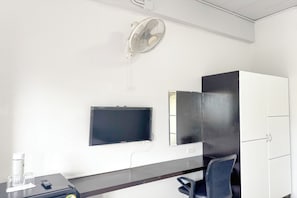 A hotel room with a desk, a chair, a TV mounted on the wall, a white cabinet, and a ceiling fan.