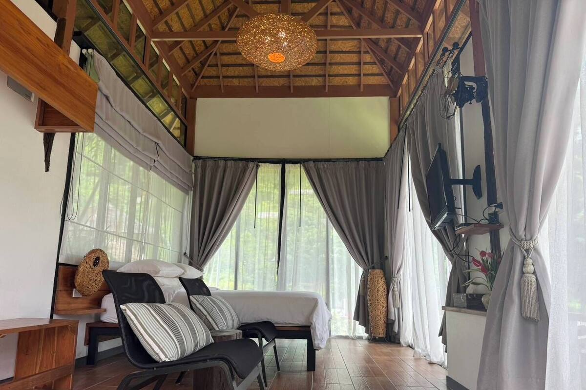 A cozy room with a bed, a chair, and a wooden bench. There are large windows with curtains, a hanging light fixture, and a shelf with decorative items.