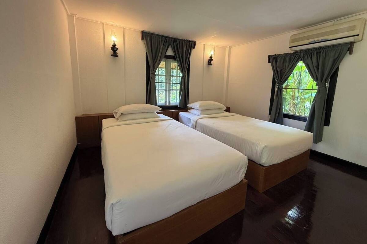 A room with two single beds, a window with curtains, an air conditioning unit, and wall-mounted lights.