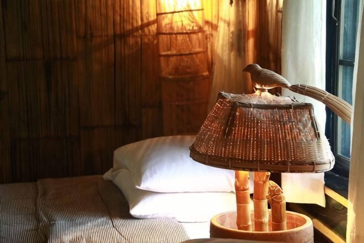 A lamp with a bird perched on it, a bed with a white pillow, and a window with a view of a bamboo structure.