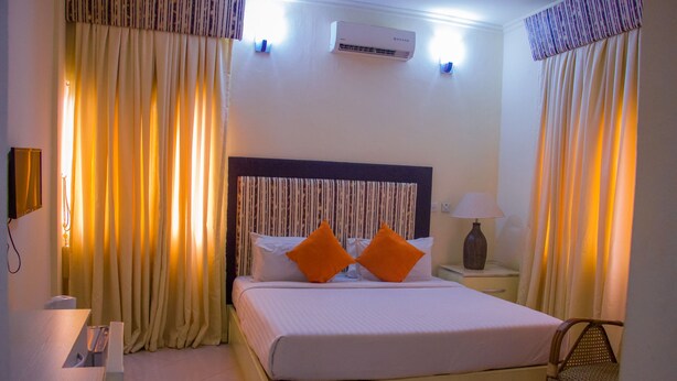 A hotel room with a bed, curtains, an air conditioner, a nightstand, and a lamp. - Pilgrims Brook Hotels Ltd (Lagos)