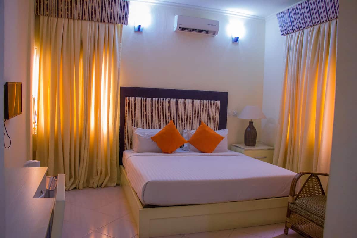 A hotel room with a bed, curtains, an air conditioner, a nightstand, and a lamp.