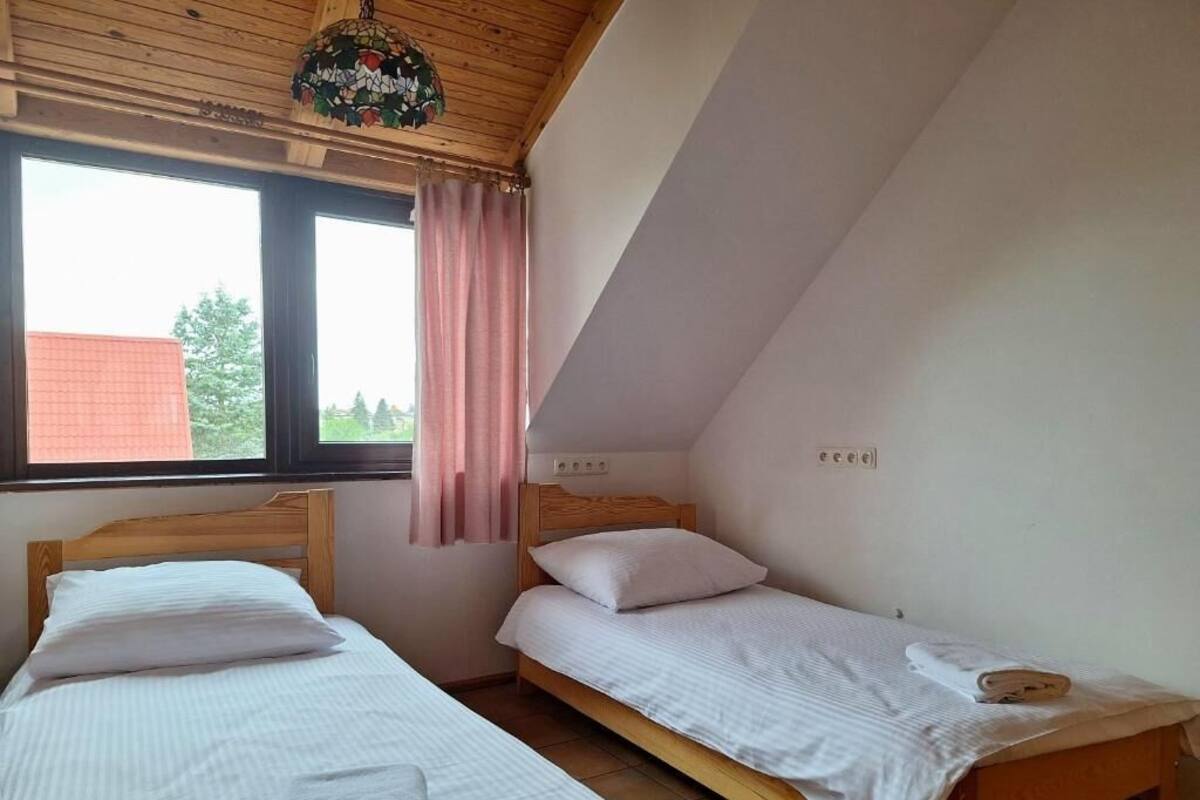 Un dormitorio con dos camas individuales, techo de madera, ventana con vista a árboles y una cortina rosa.