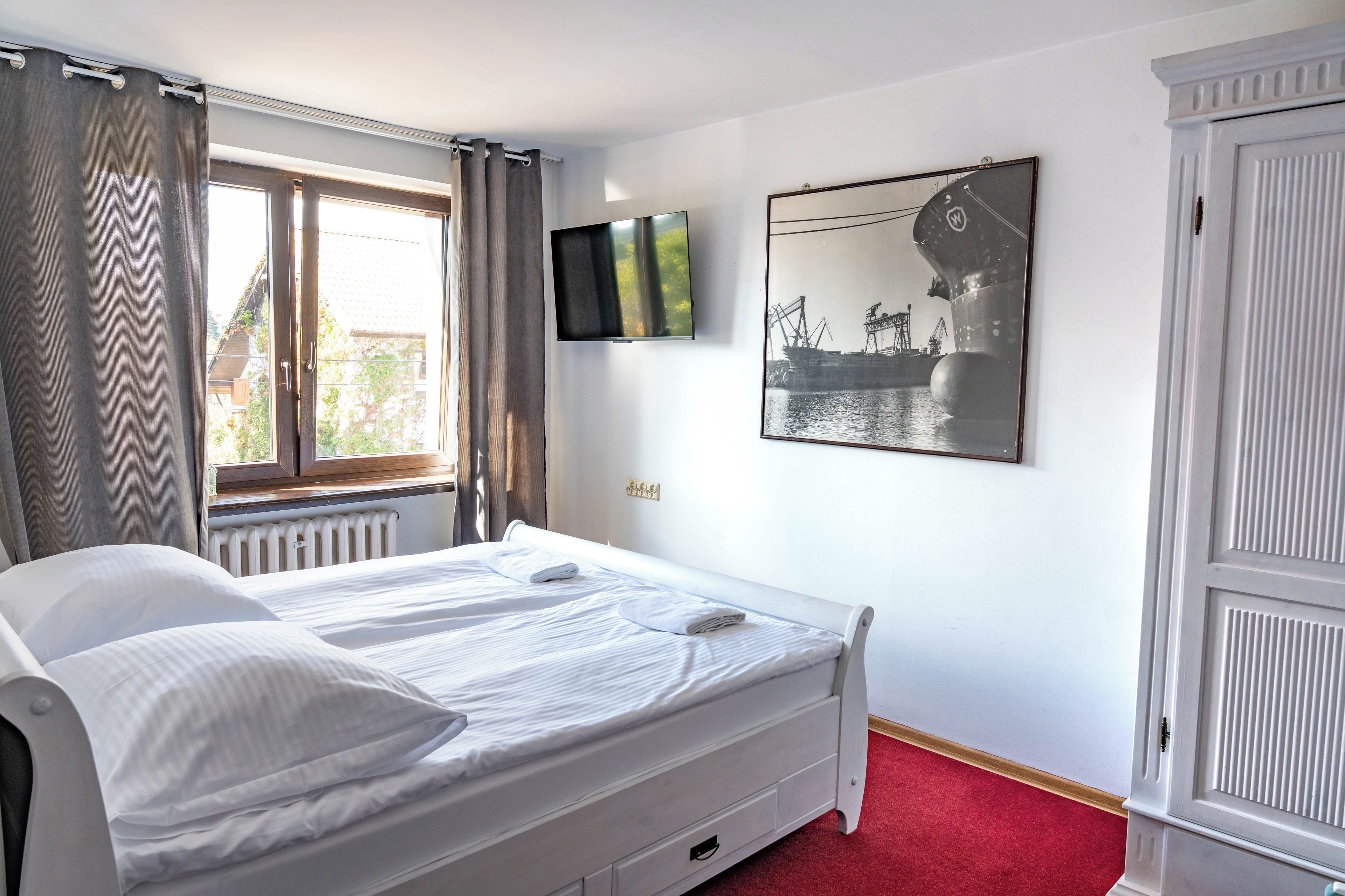 A bedroom with a bed, a TV, a window with curtains, a radiator, and a door.