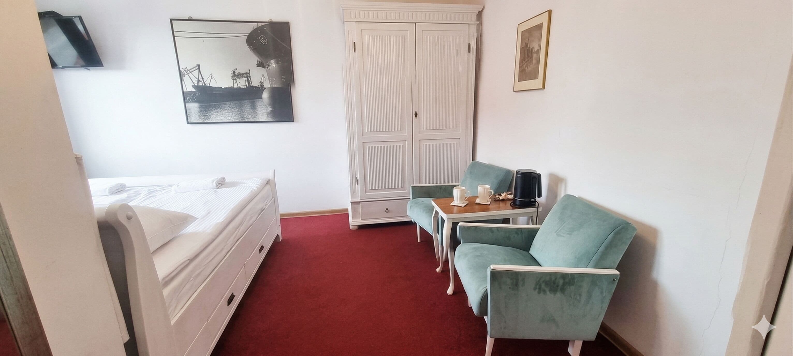 A hotel room with a bed, a desk, a chair, a wardrobe, and a painting on the wall.