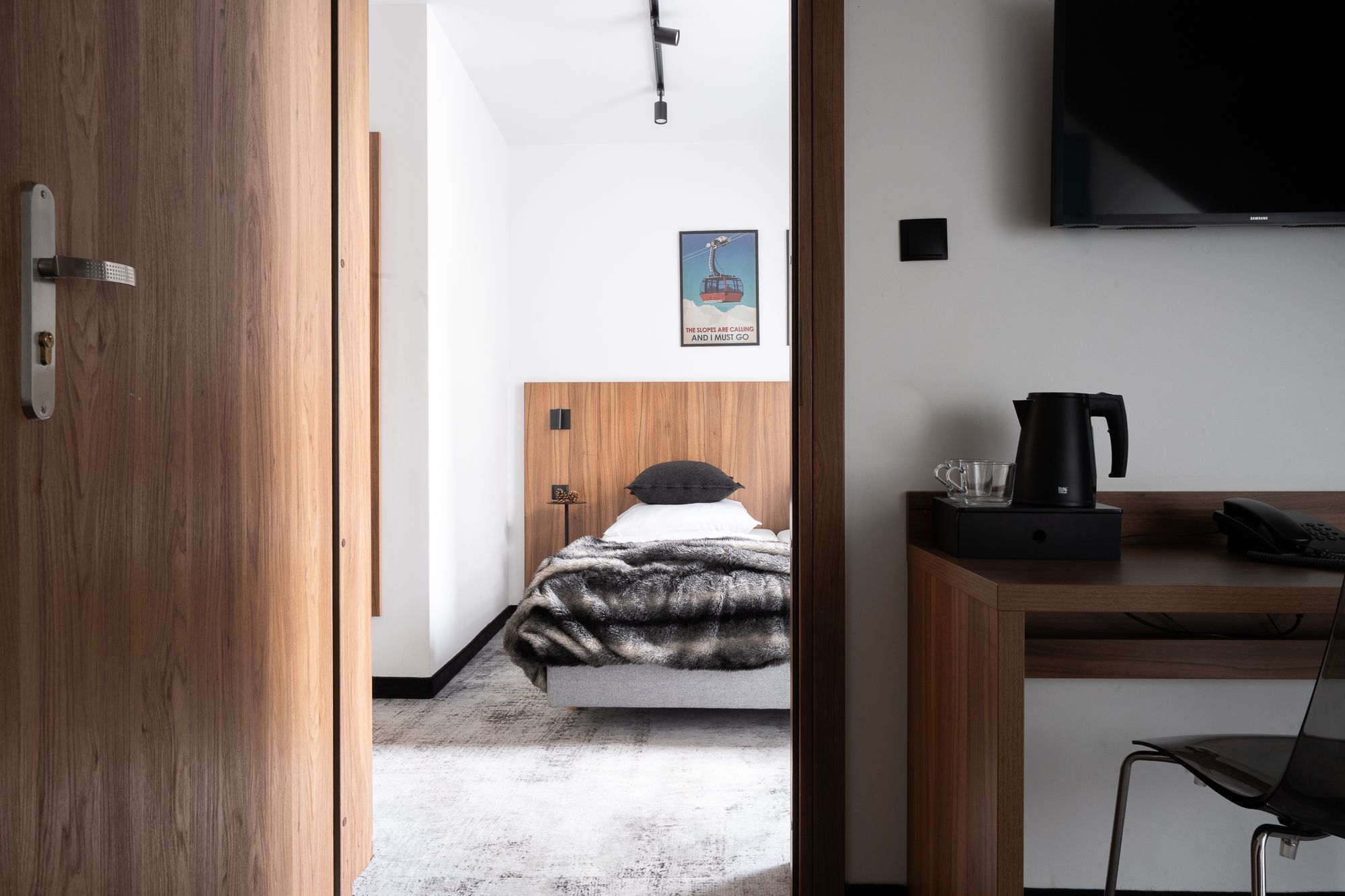 Ein Hotelzimmer mit Holztür, einem Bett mit schwarz-weiß gestreifter Decke, einem Wand-Fernseher, einer Kaffeemaschine und einem Schreibtisch mit Stuhl.