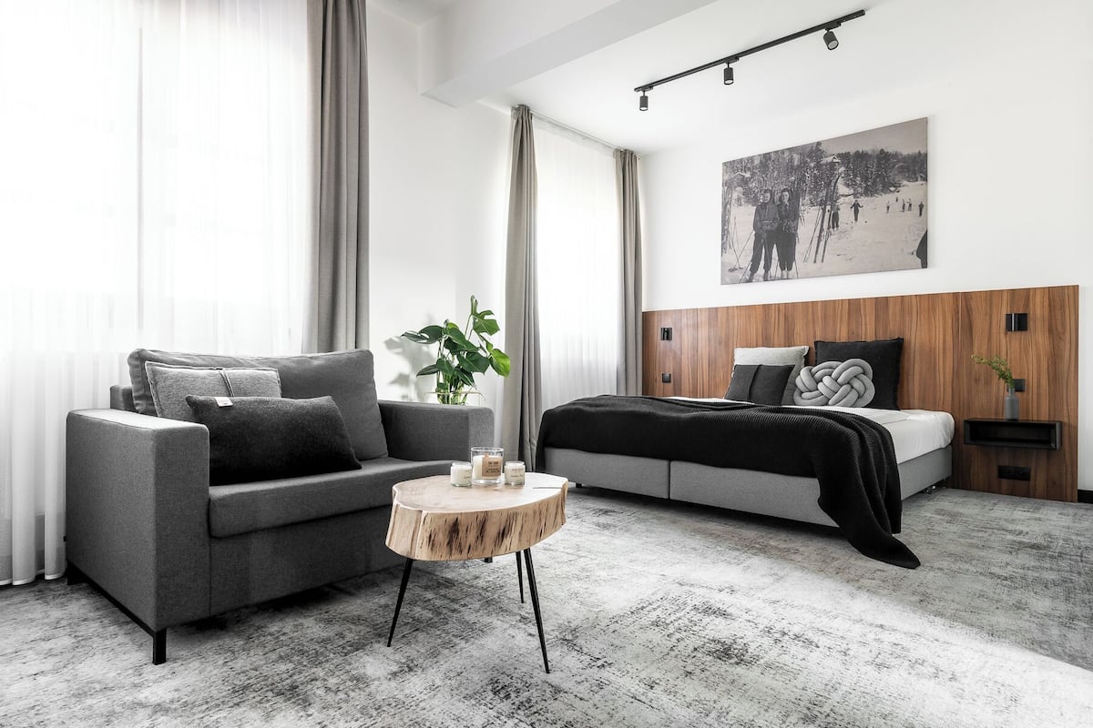A modern bedroom with a grey sofa, a round wooden side table, a bed with a wooden headboard, and a large framed picture on the wall.