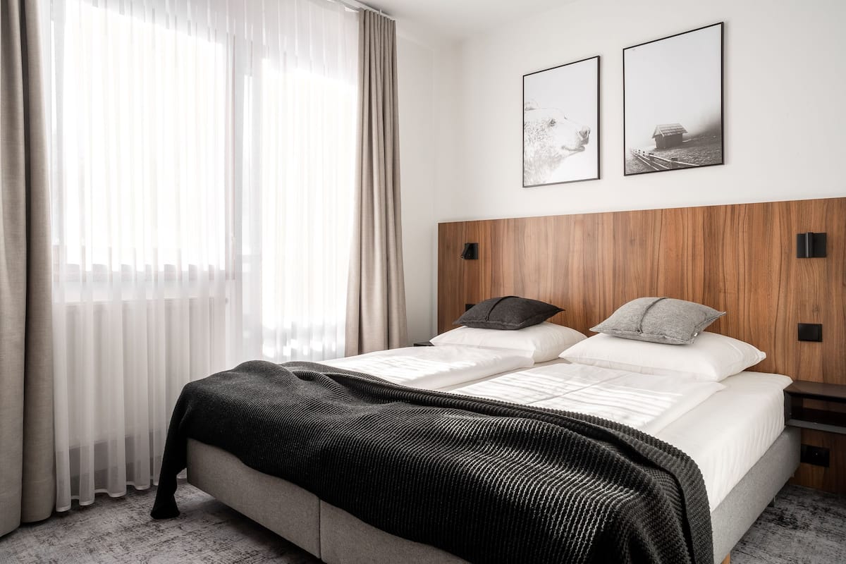 A modern hotel room with a bed, a wooden headboard, two framed pictures on the wall, and a window with sheer curtains.