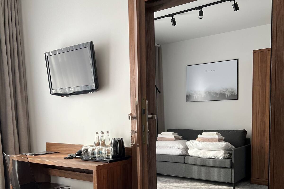 A hotel room with a wooden desk, a bed with a black headboard, a grey sofa, a TV mounted on the wall, and a painting above the sofa.