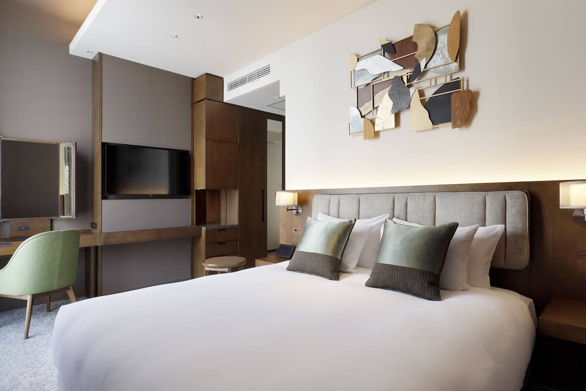 Quarto de hotel com cama, escrivaninha, cadeira, televisão e uma obra de arte na parede.