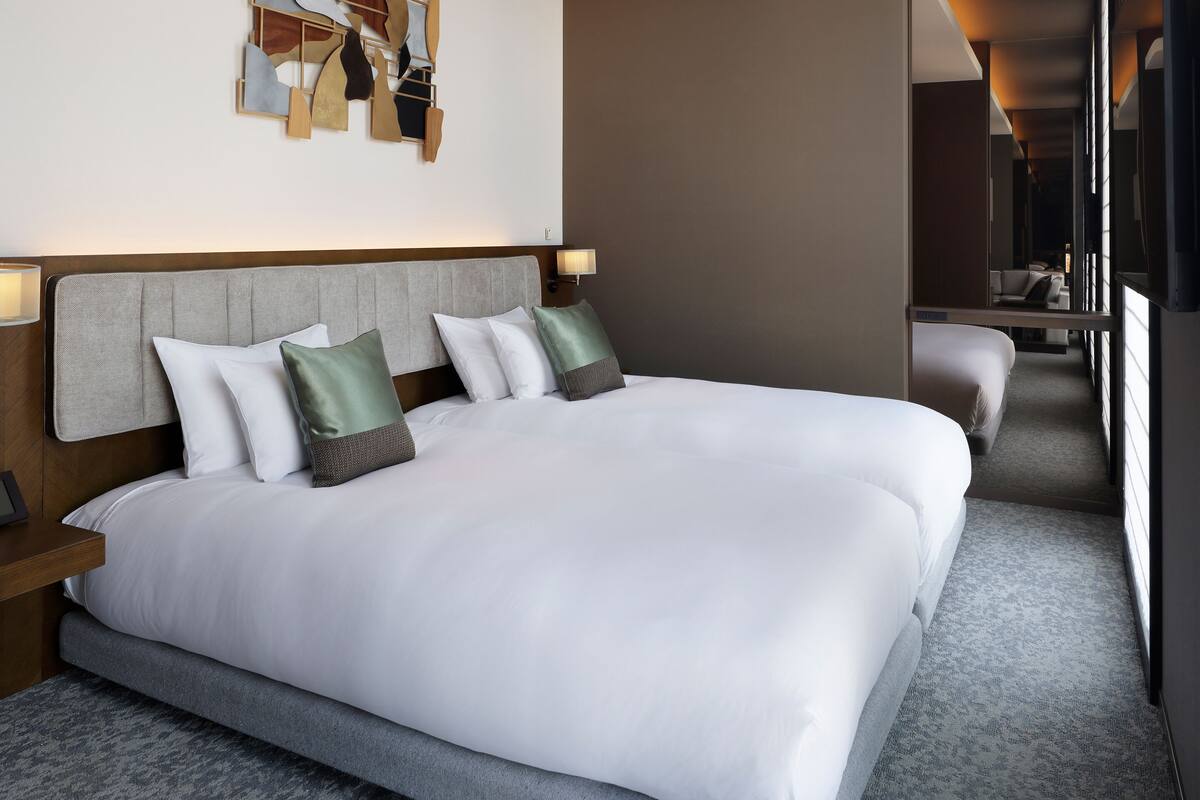 Quarto de hotel com uma cama grande, cabeceira, mesas de cabeceira, uma obra de arte na parede e vista para outro quarto.