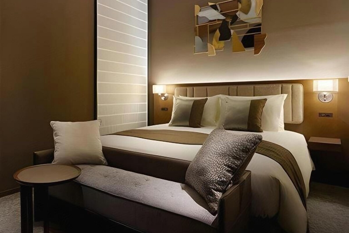 A modern hotel room with a large bed, a bedside table, a bench, and a wall-mounted abstract art piece.