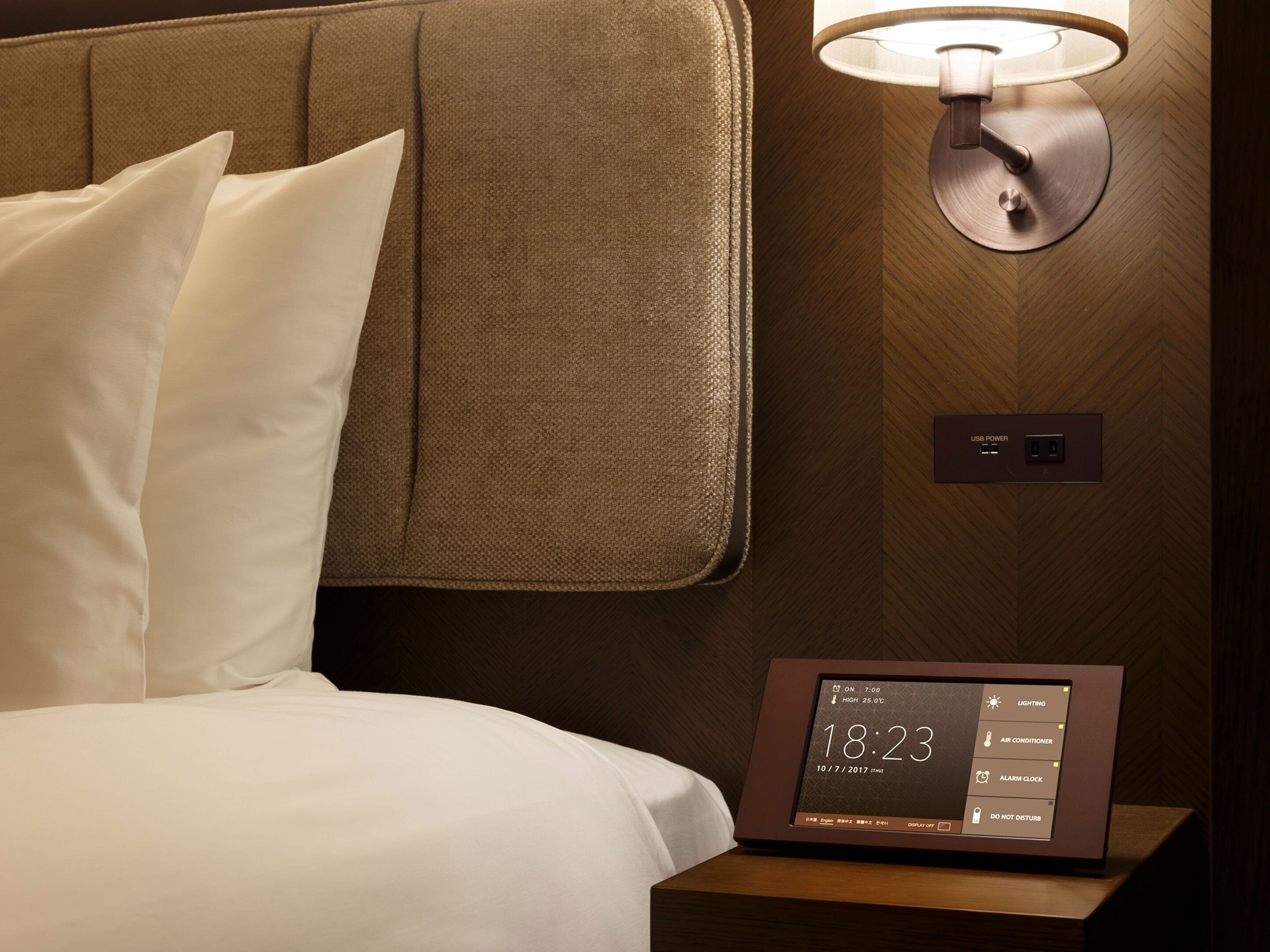 A hotel room with a bed, pillows, a nightstand with a digital clock, a wall-mounted lamp, and a USB power outlet.