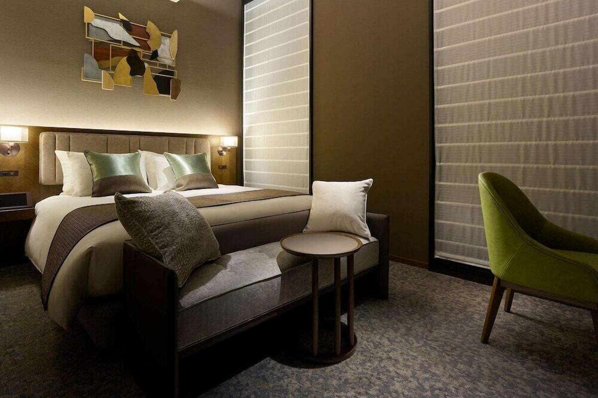 Quarto de hotel moderno com uma cama, uma pequena mesa redonda, uma poltrona verde e uma grande obra de arte abstrata na parede.