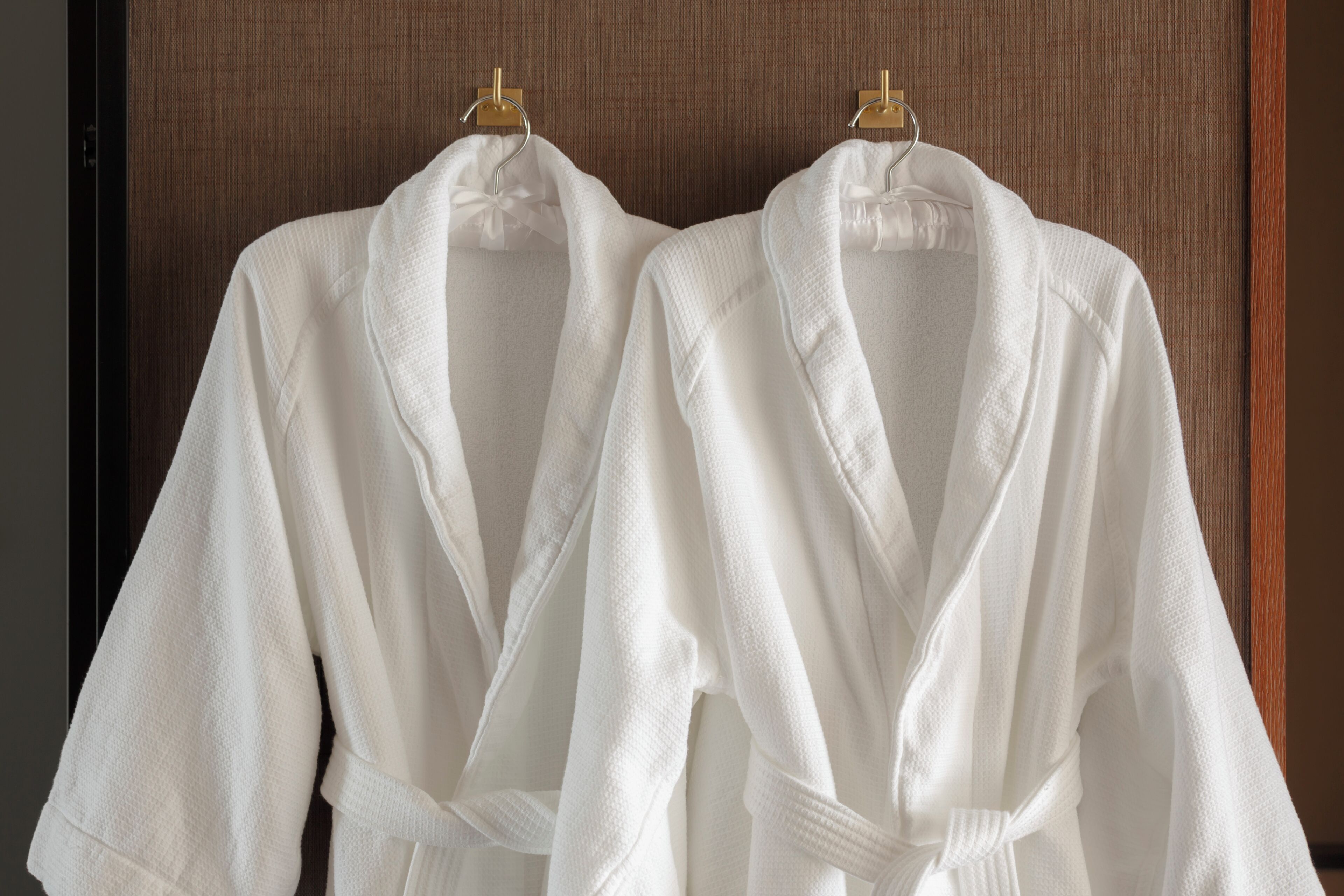 Two white bathrobes hanging on wooden hangers against a textured wall.