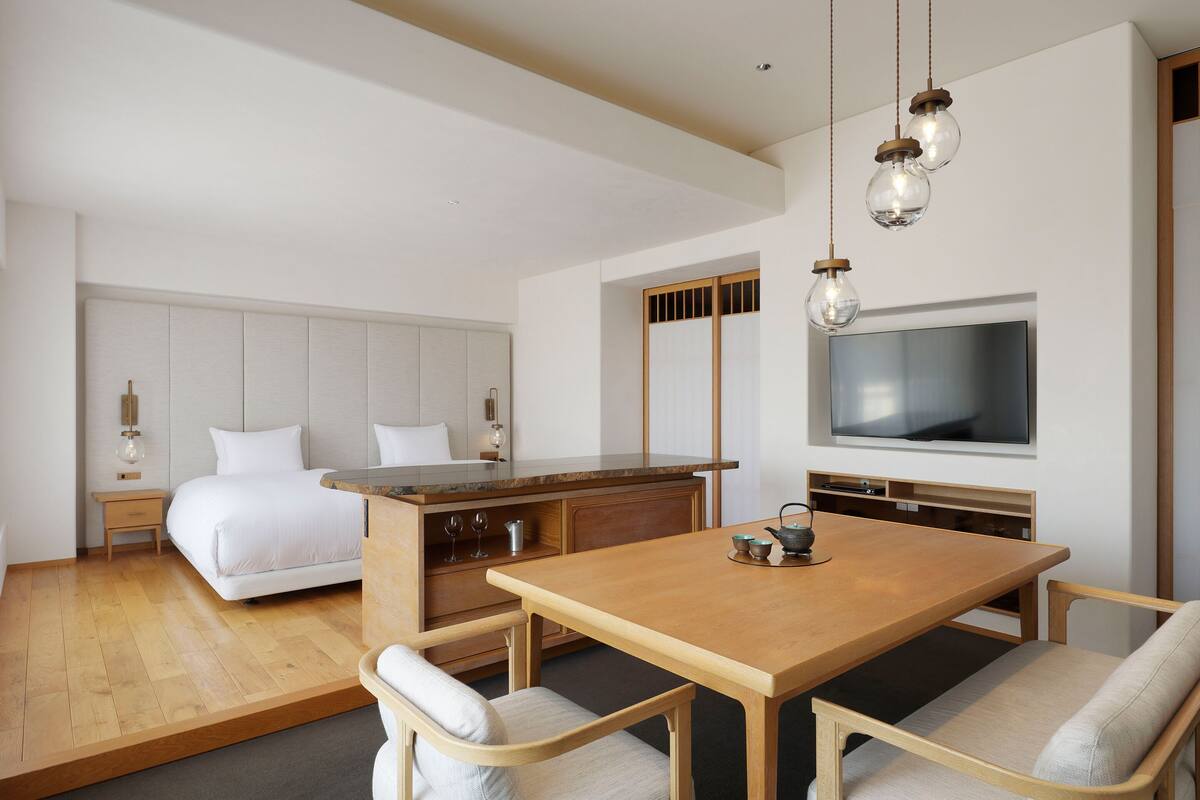 A modern hotel room with a bed, a desk, a TV, and a dining area with chairs.