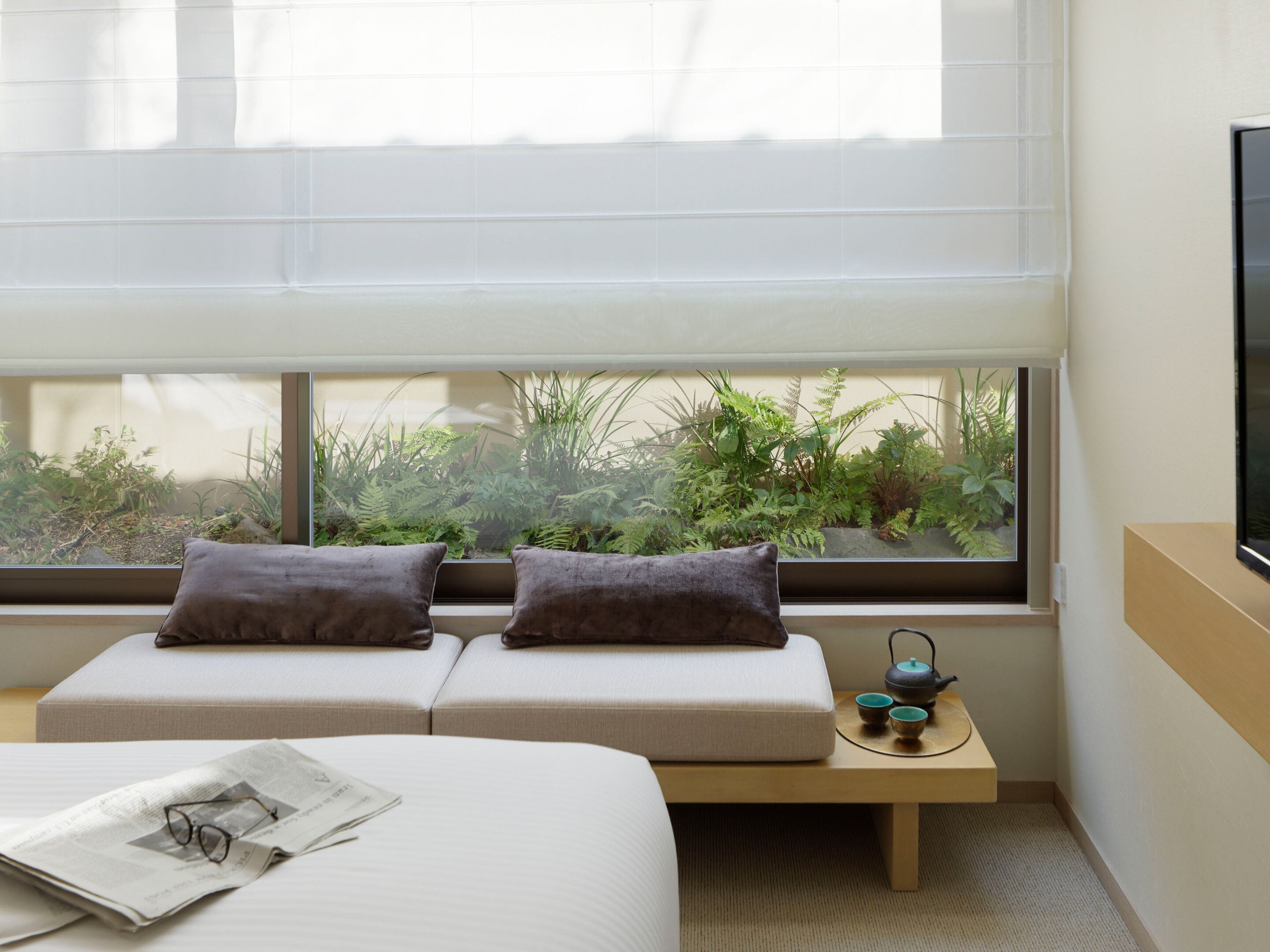 Ein modernes Zimmer mit einem Bett, zwei gepolsterten Sitzgelegenheiten, einem kleinen Tisch mit Teekanne und Blick auf Grün hinter einem großen Fenster.