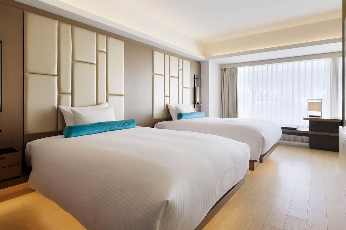 A hotel room with two beds, a large window with curtains, a bedside table, and a wall-mounted light fixture.