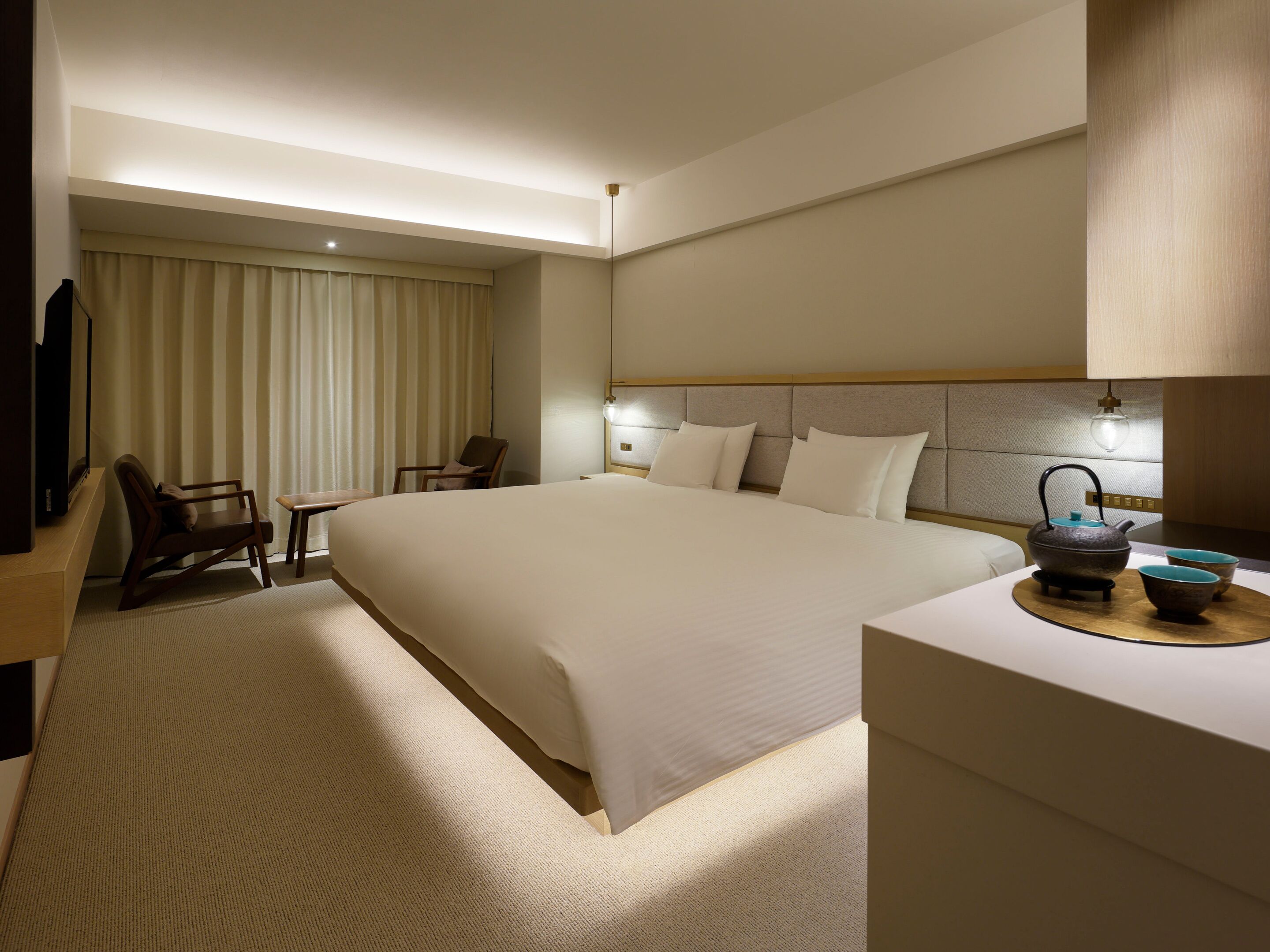 A hotel room with a large bed, a desk, a chair, a television, and a tea set.