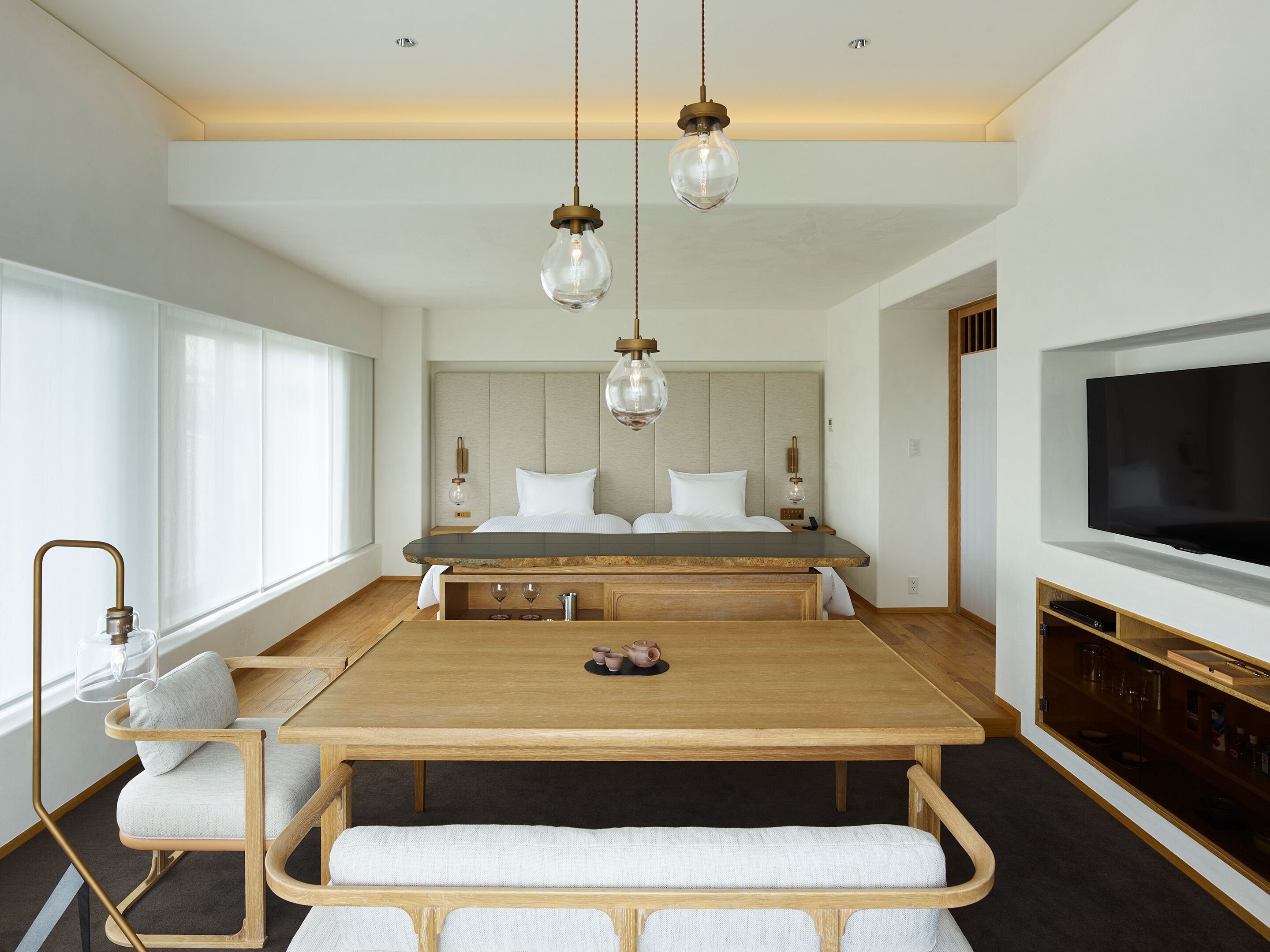 Quarto moderno com cama grande, mesas de cabeceira, televisão e área de refeições com mesa e cadeiras de madeira.