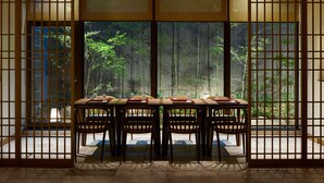 Quarto tradicional japonês com piso de esteira de tatame, uma mesa baixa com quatro cadeiras e grandes portas deslizantes com vista para bambus lá fora.