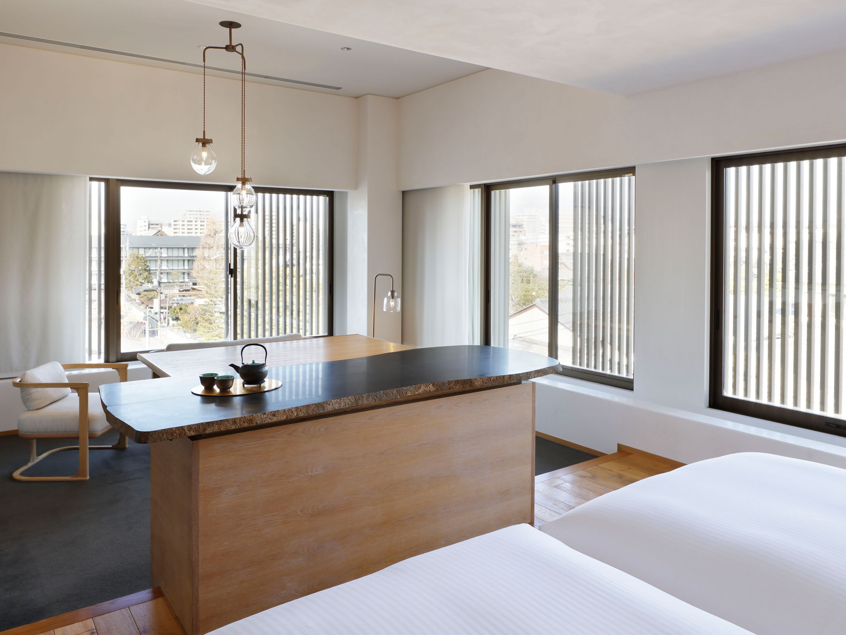 Una habitación de hotel moderna con una cama grande, un escritorio de madera, una silla y ventanas amplias con persianas.