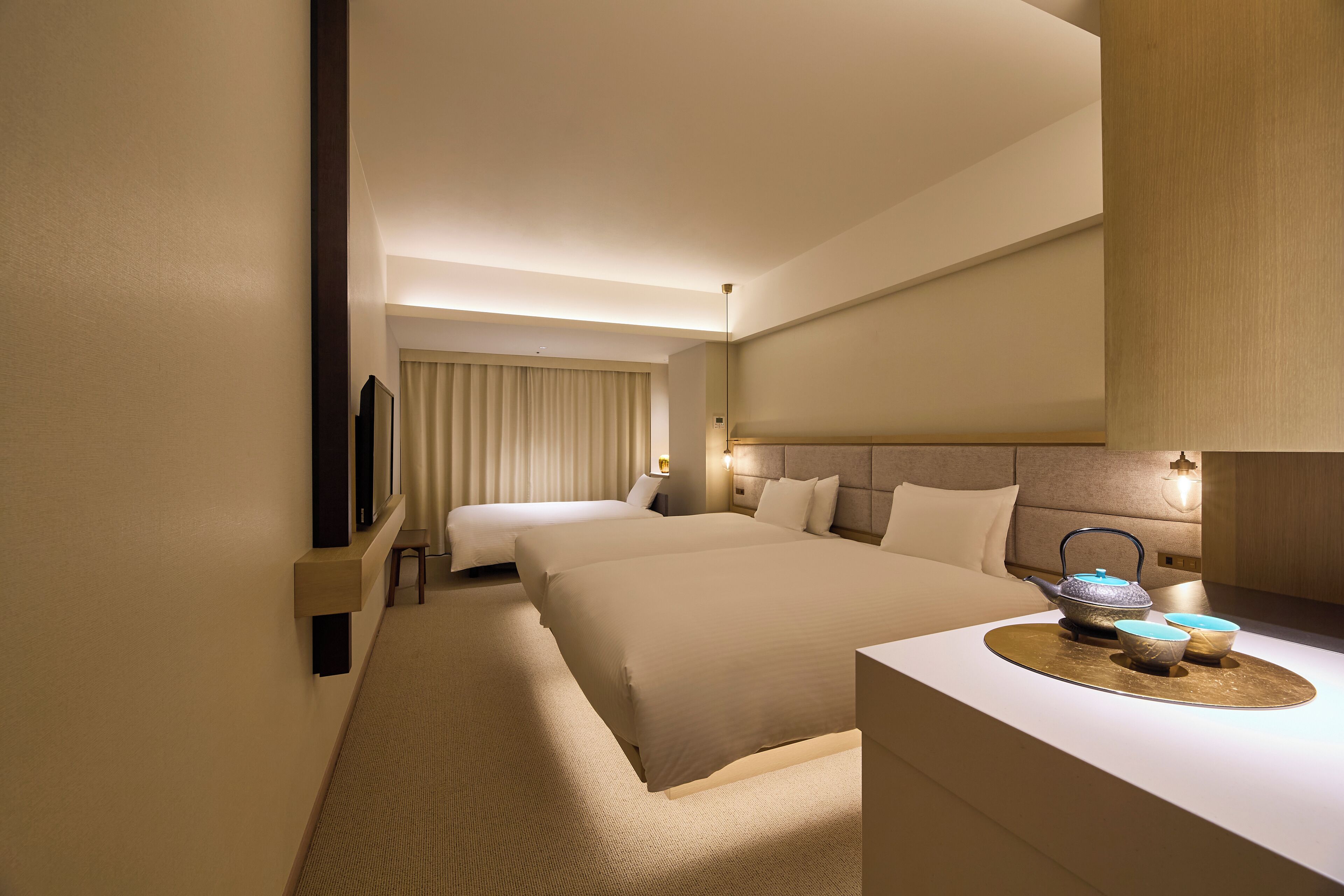 Habitación de hotel con dos camas, un televisor de pantalla plana, una mesita de noche con lámpara y una bandeja decorativa con tetera y tazas.