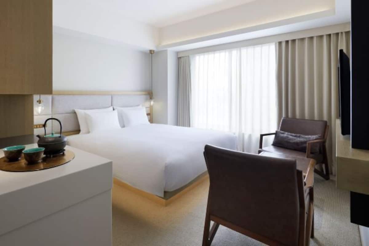 Habitación de hotel con una cama grande, un escritorio con silla, televisor y ventana con cortinas.