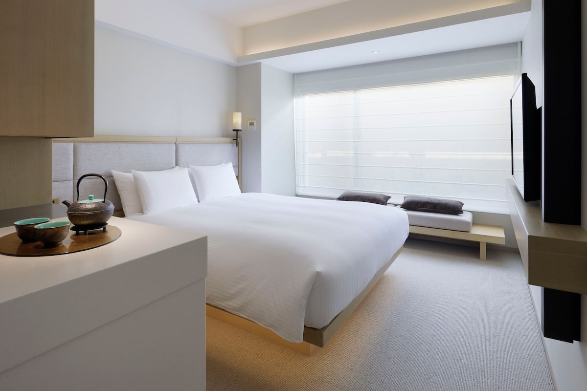 A hotel room with a bed, a bedside table, a TV, and a window with blinds.