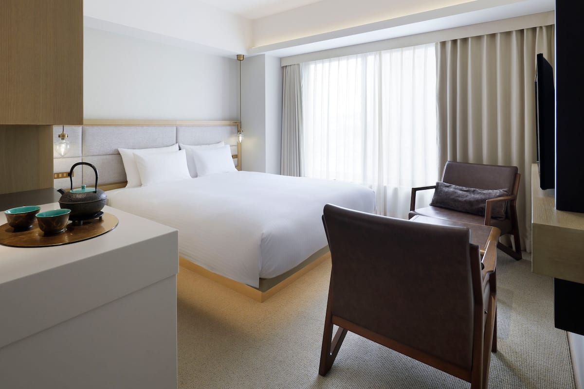 A hotel room with a bed, a chair, a desk with a kettle and cups, and a television.