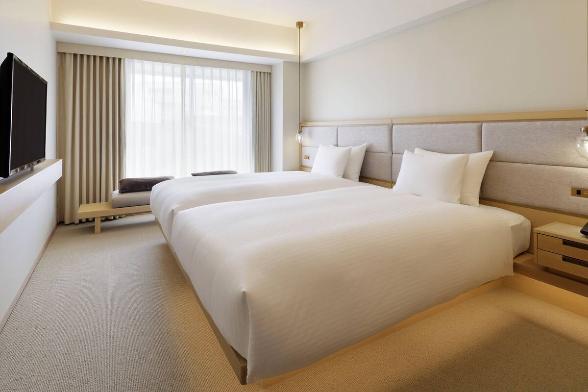 A hotel room with two beds, a TV, a sofa, and a large window with curtains.