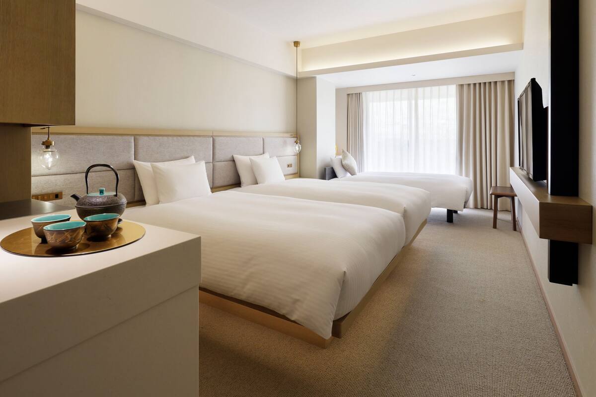 A hotel room with two beds, a desk with a kettle, cups, and a flat-screen TV.