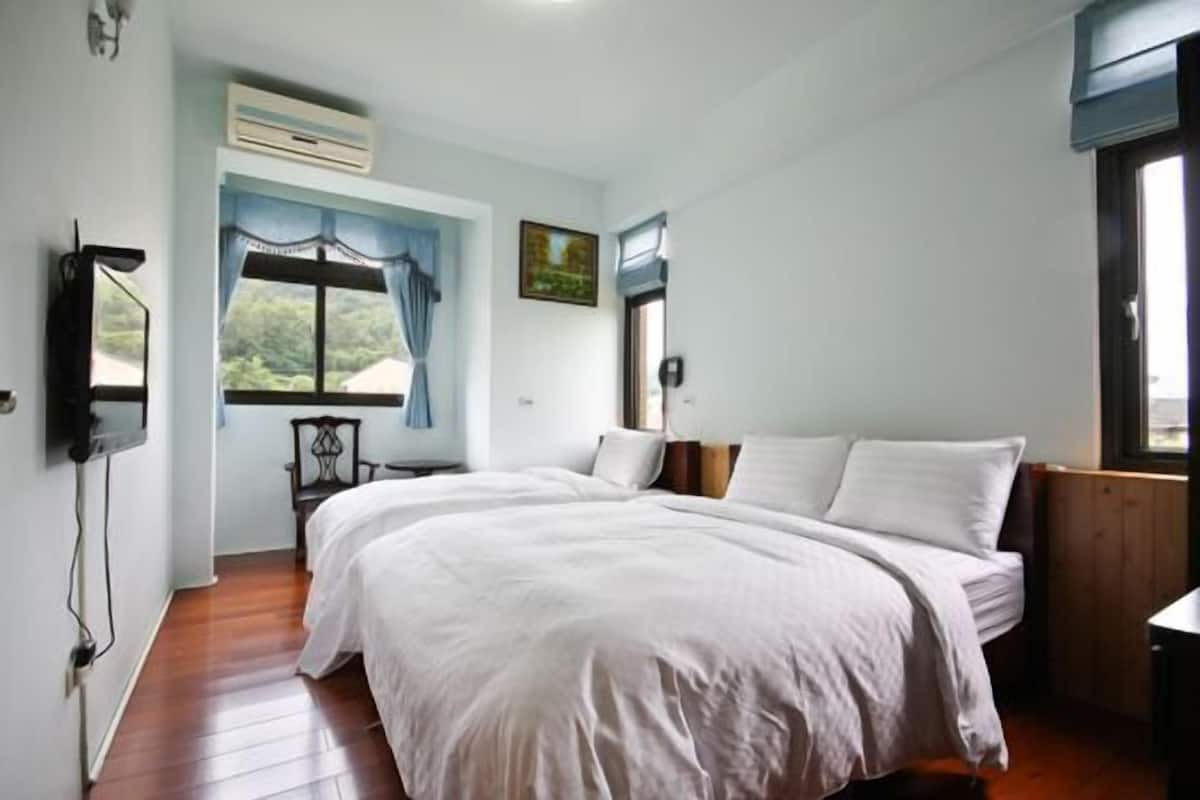 Quarto com uma cama grande, janela com cortinas, uma cadeira e TV fixada na parede.