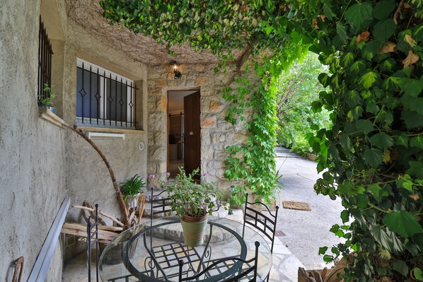 A stone building with a circular outdoor seating area, a window with bars, and a door leading inside. - Le Mazet de Tourrettes (Tourrettes-sur-Loup)