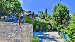 A stone wall with a potted plant in the foreground, a metal gate, and a building with a tiled roof and greenery in the background. - Le Mazet de Tourrettes (Tourrettes-sur-Loup)