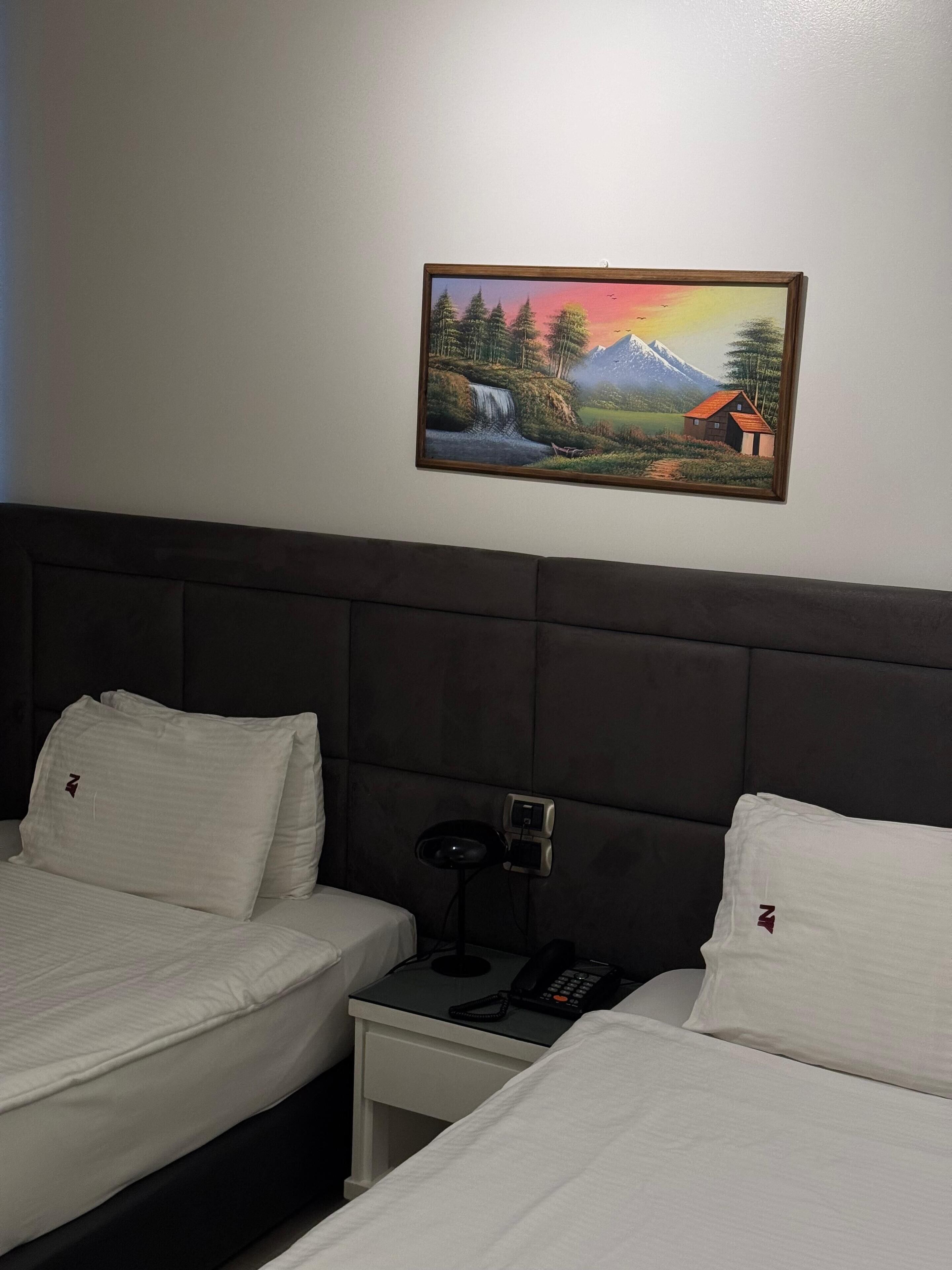 A hotel room with two beds, a nightstand, and a painting of a mountainous landscape.