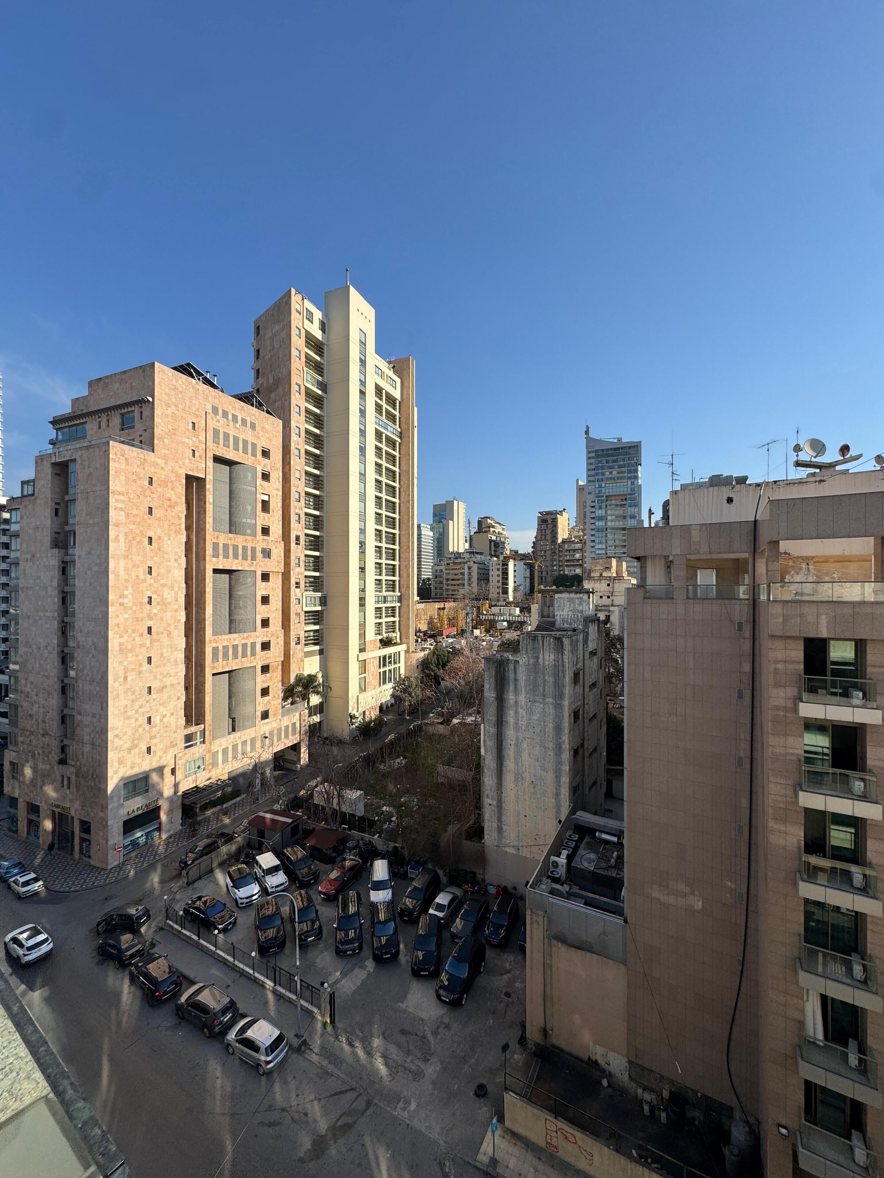 A high-rise building with multiple floors, a parking lot with various cars, and other surrounding buildings.