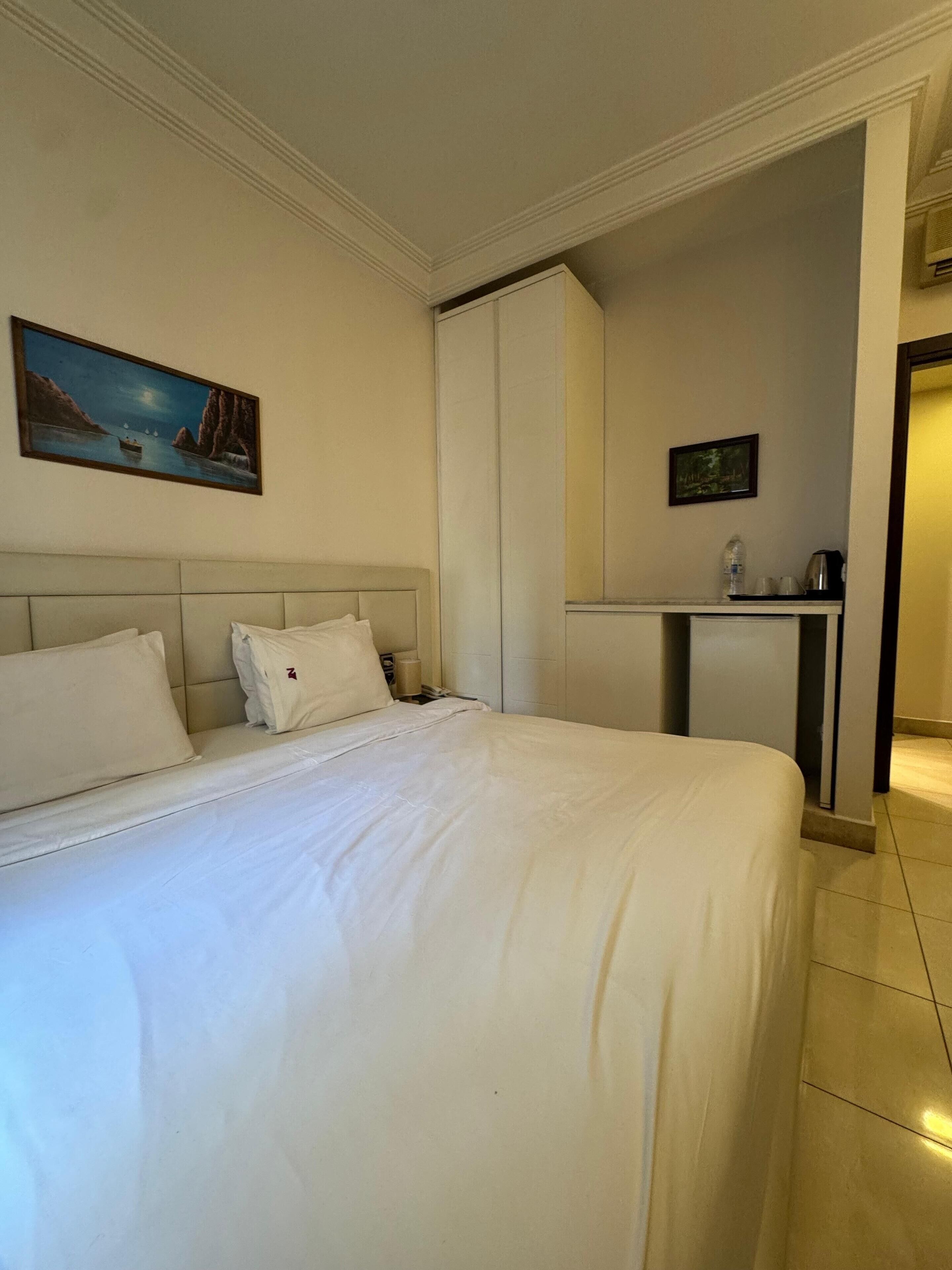 A hotel room with a large bed, a painting of a coastal scene, a wardrobe, and a small kitchenette area.