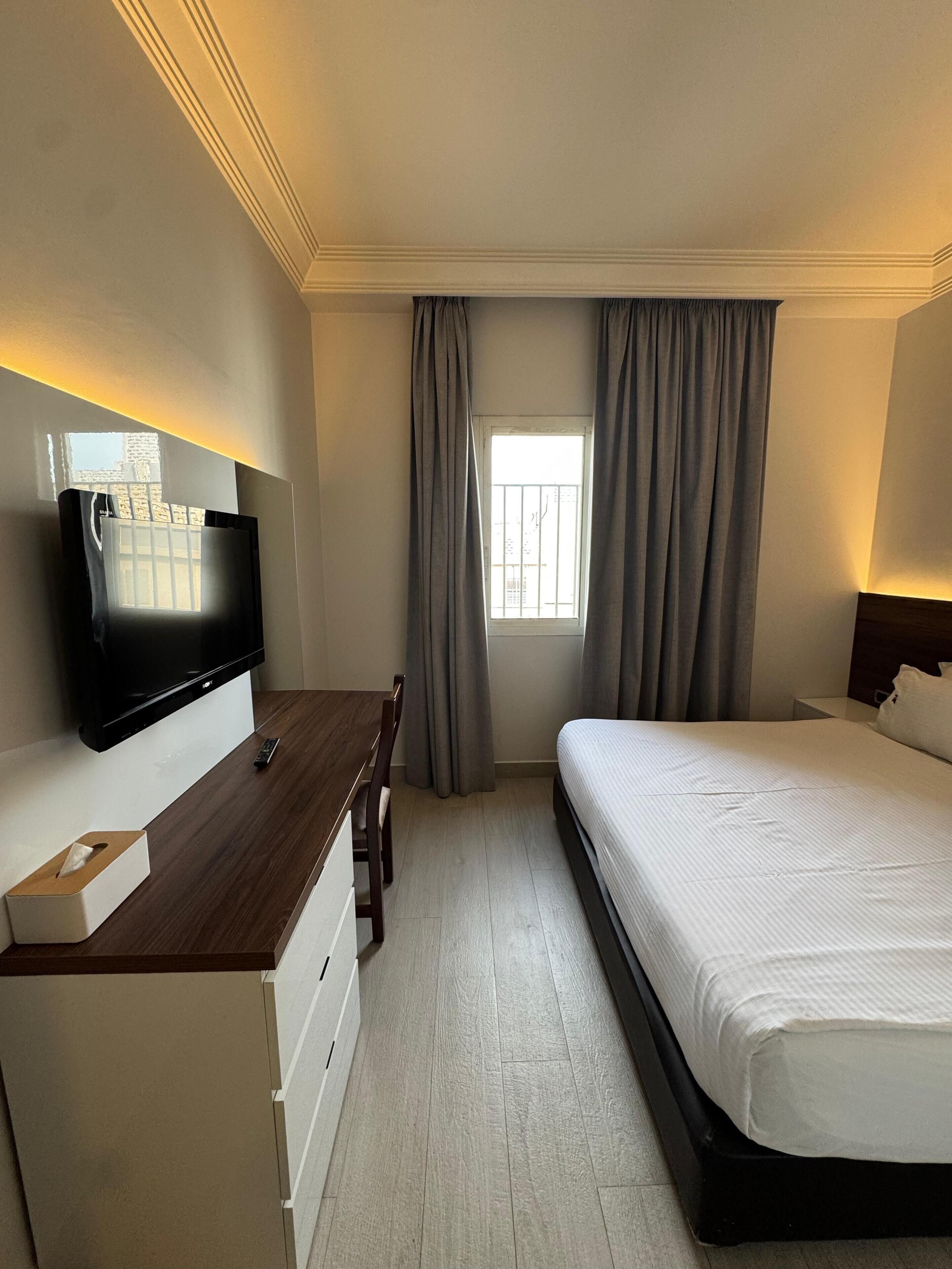 Ein Hotelzimmer mit Bett, Schreibtisch mit Fernseher, Fenster mit Vorhängen und Nachttisch.