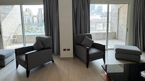 Una sala de estar moderna con sofás grises, una mesa de centro de madera y vistas a un paisaje urbano a través de amplios ventanales.