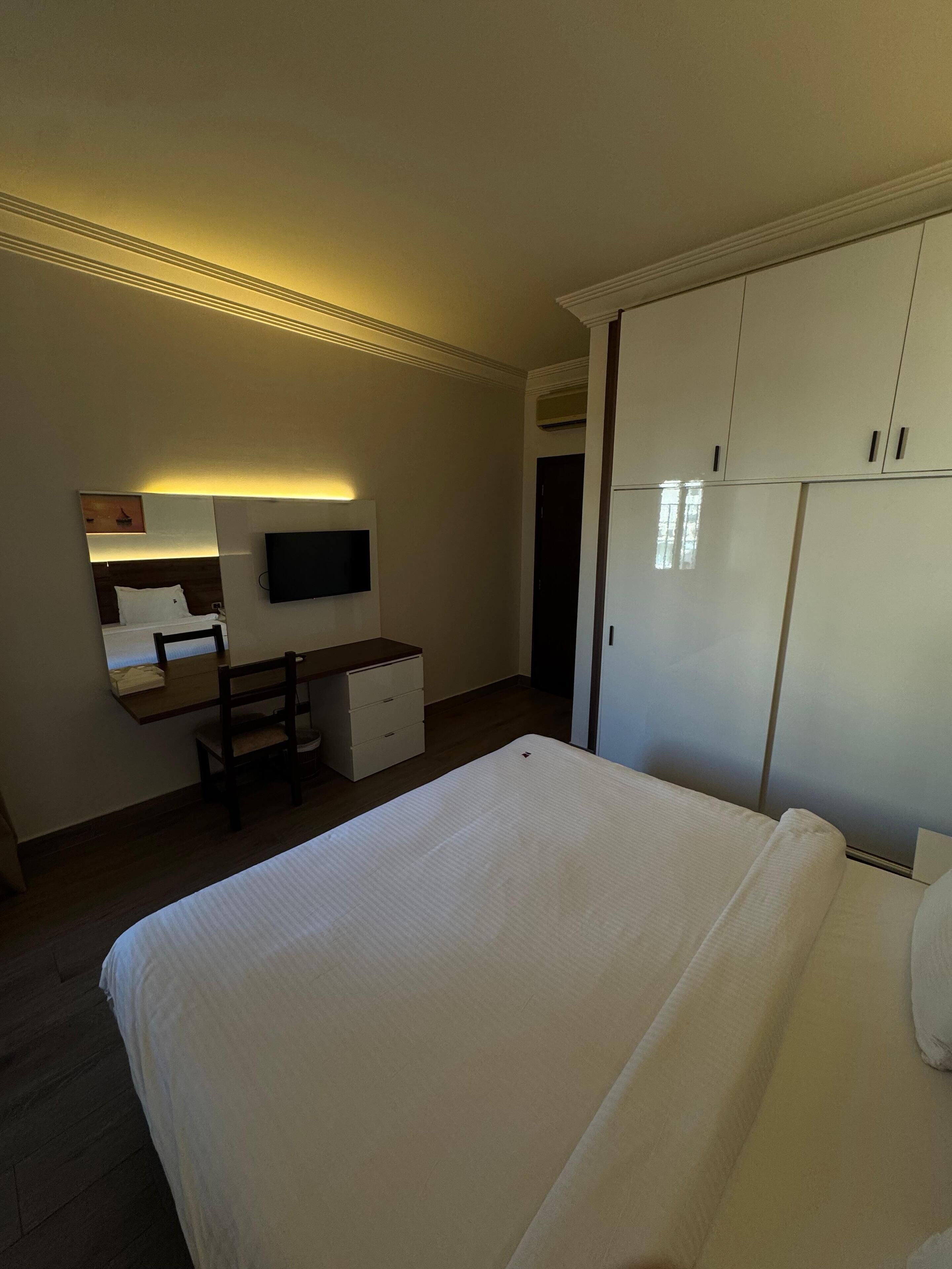 Et hotelværelse med seng, skrivebord med stol, fjernsyn og garderobeskab.