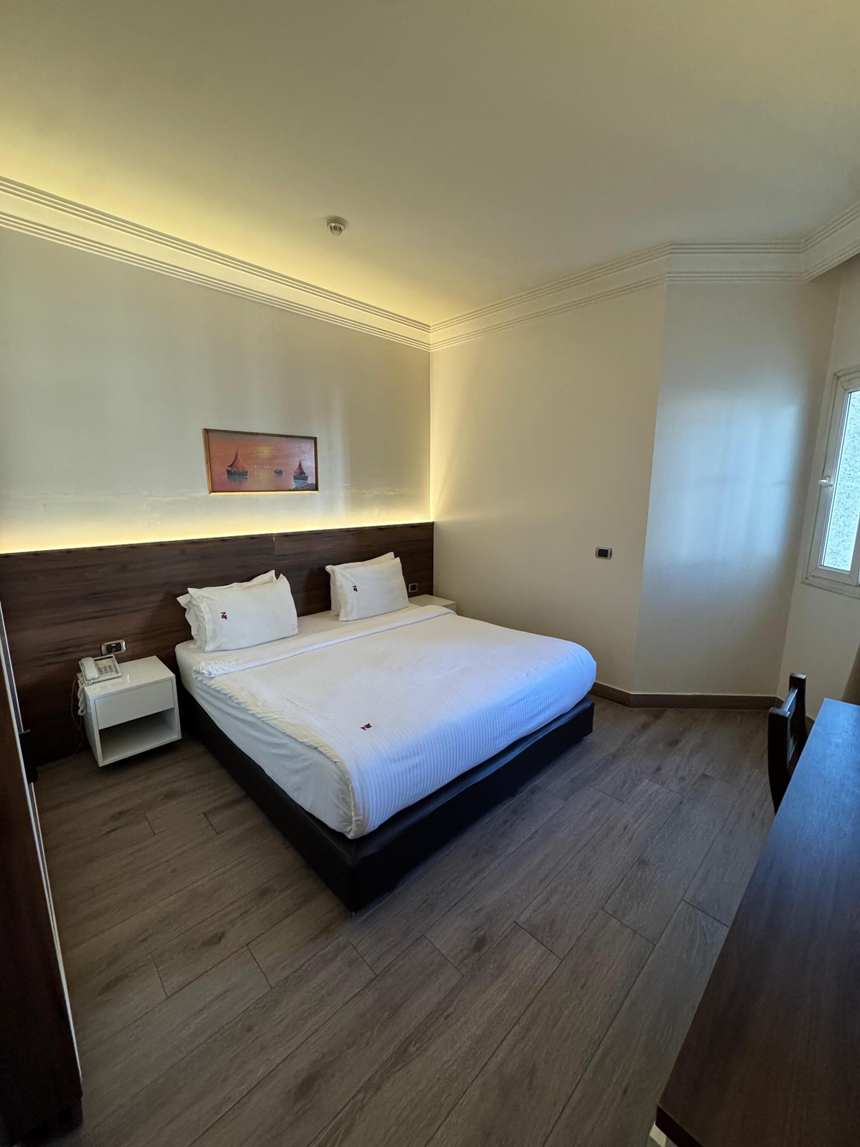 Una habitación de hotel con una cama grande, una mesita de noche con un teléfono, una lámpara de noche y un cuadro en la pared.