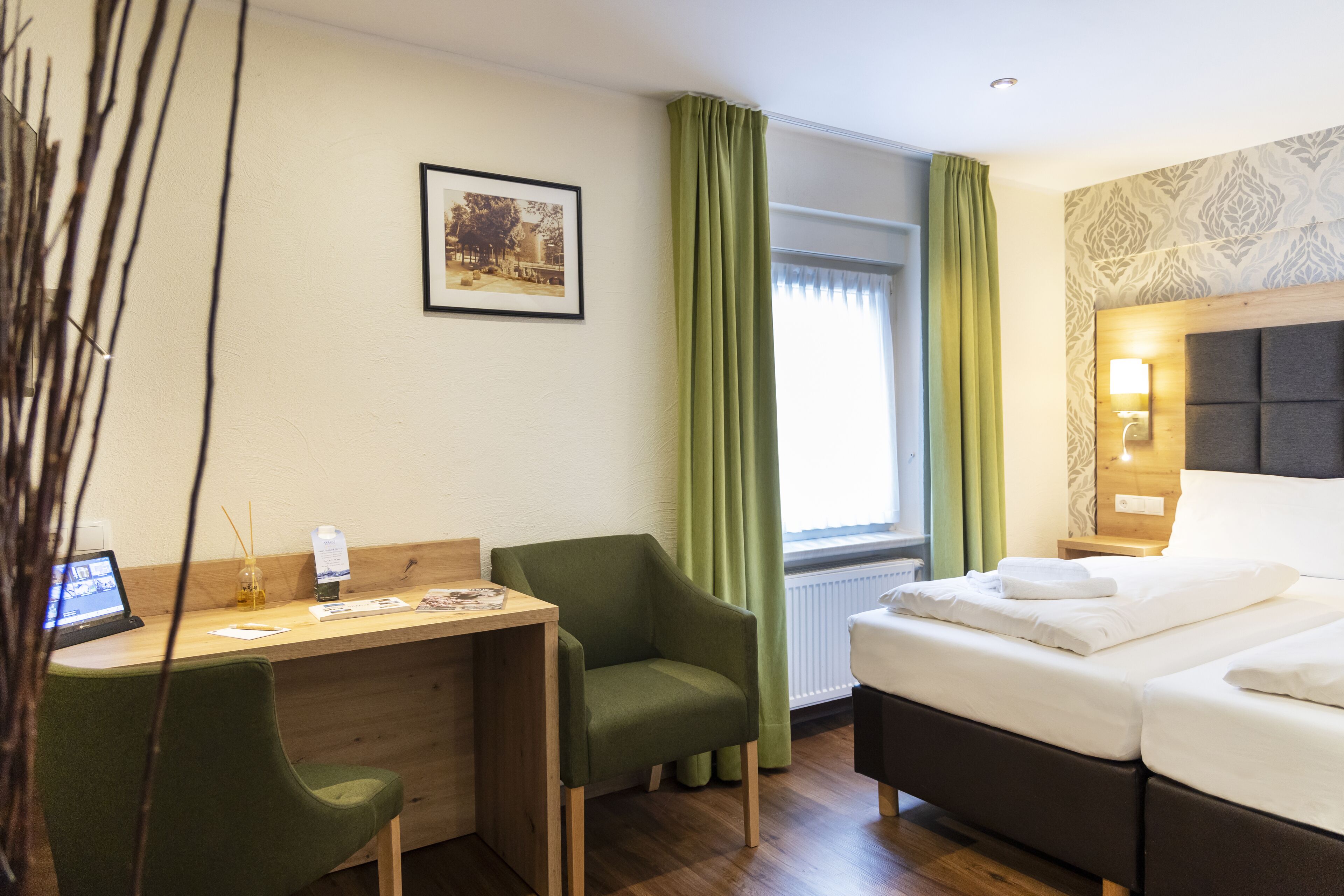 Habitación de hotel con cama, escritorio, dos sillas, un cuadro en la pared y una ventana con cortinas.