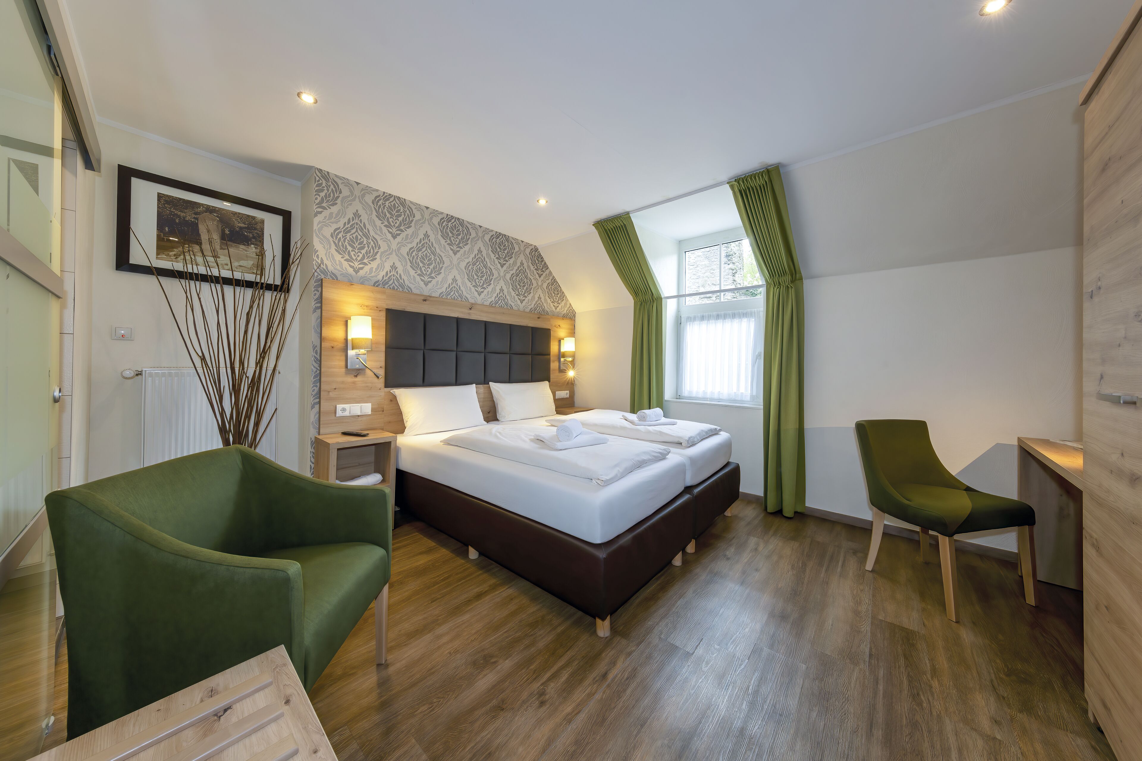 Une chambre d’hôtel moderne avec un grand lit, deux tables de chevet, un fauteuil vert et un bureau en bois.