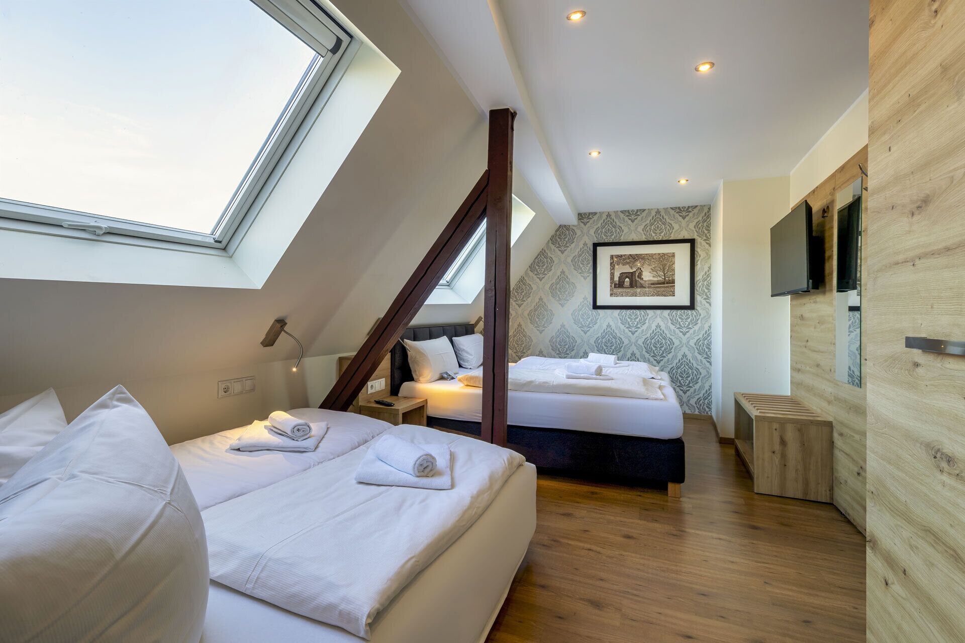 A modern bedroom with a skylight, a large bed, a bedside table, a wooden bench, and a wall-mounted TV.