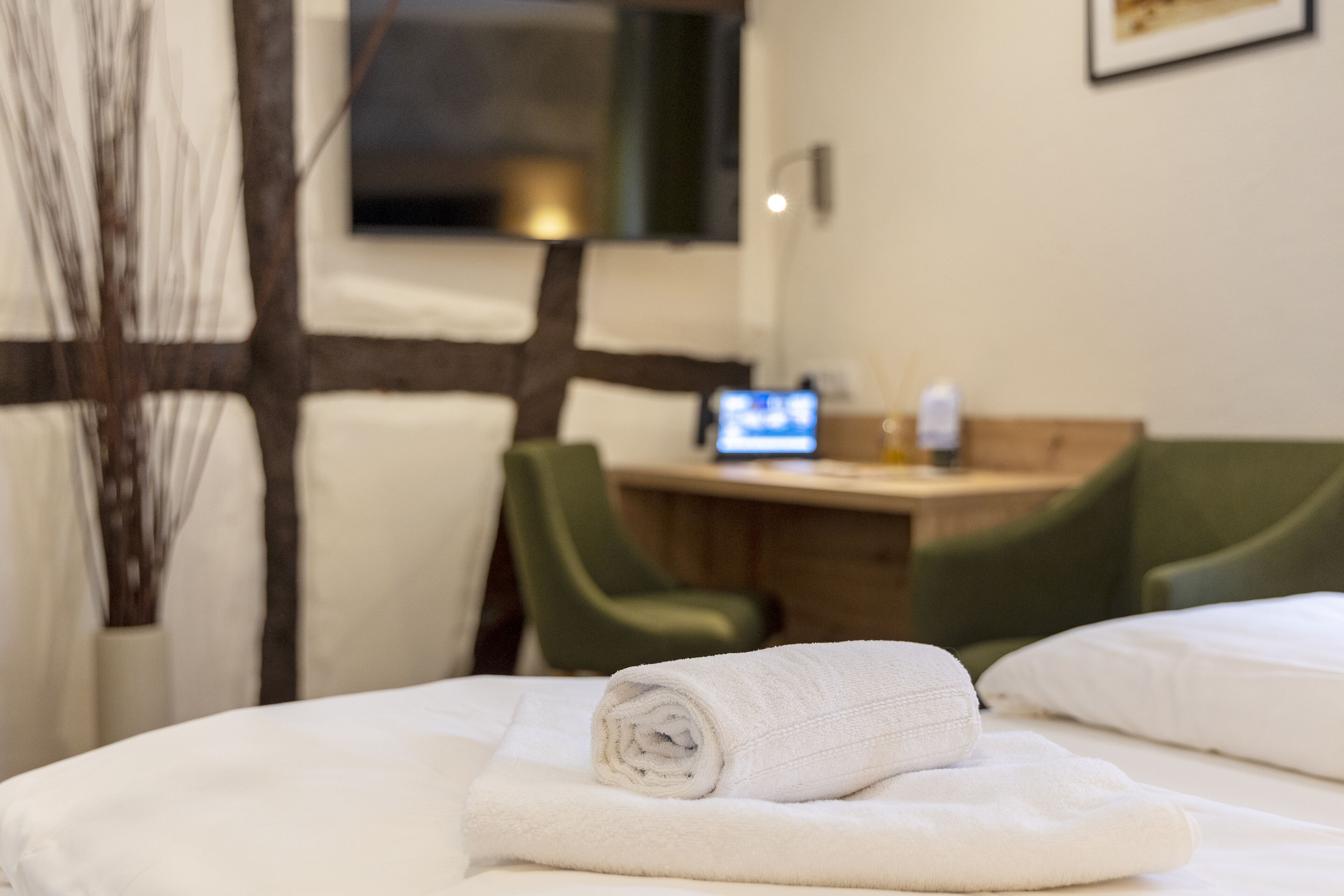 A neatly made bed with white linens, a small desk with a computer, and green chairs in a hotel room.