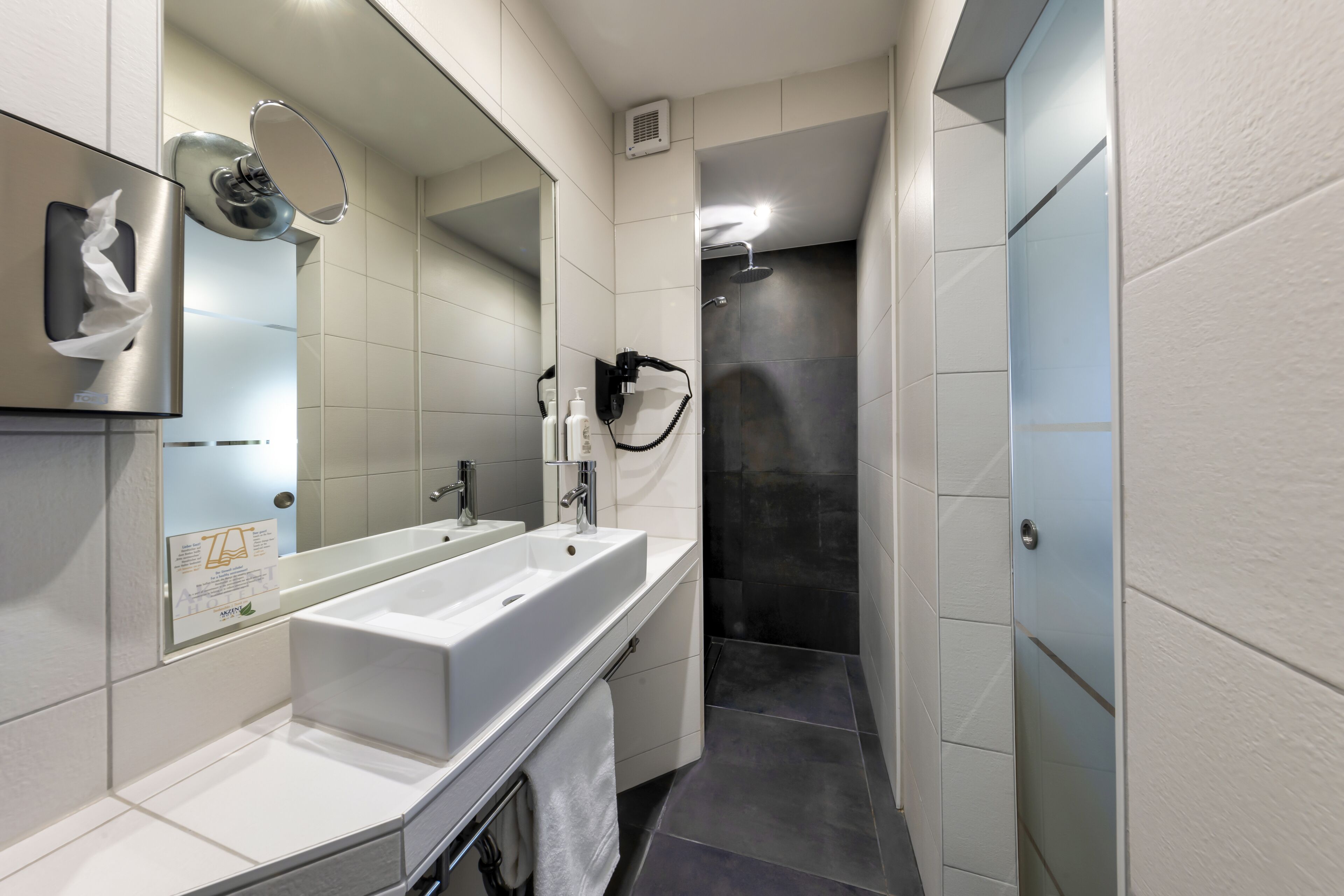 A modern bathroom with a large mirror, two sinks, a towel rack, and a shower area.